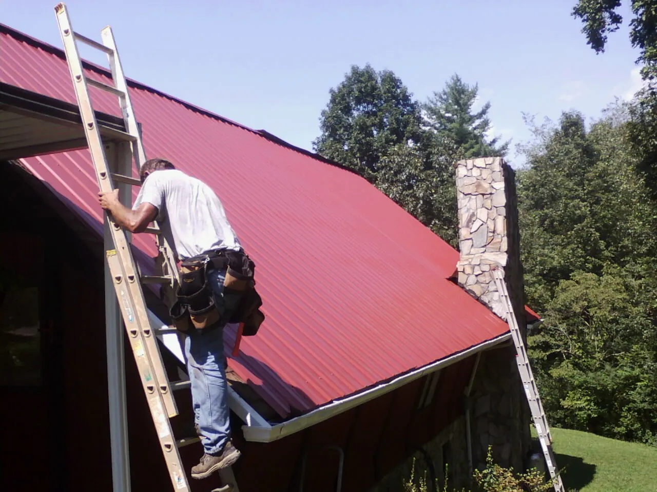 Roofer on a ladder beside a red metal roof and stone chimney — Soffit & Fascia Repair by Roofing Brothers in Pendleton